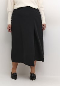 Zwarte rok tot halverwege met een asymmetrische zoom en een gladde stof. Gecombineerd met puntige zwarte loafers en een crèmekleurige, gestructureerde trui.