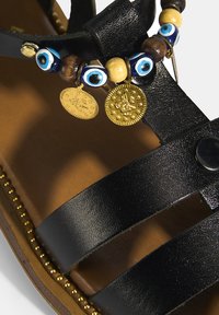 Schwarze Leder-Sandaletten mit mehreren Riemen, dekorativen Perlenakzenten in Blau, Gold und Holz, die münzenähnliche Anhänger und goldene Verzierungen aufweisen.