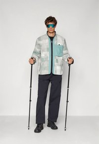 Helehall jope mintrohelise taskuga, millel on ees tõmblukk ja plokkmustrid, sobitatud tumesiniste pükstega ja mustade jalanõudega, hoiab matkakeppe.