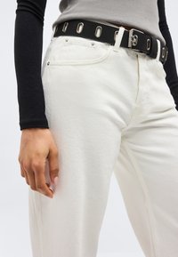 Άτομο που φοράει λευκό παντελόνι με μαύρη ζώνη που διαθέτει μεταλλικές τρύπες, γκρι μπλουζάκι μεσαίου μήκους στημένο μέσα στο παντελόνι και ένα μακρύ μαύρο μανίκι ορατό σε ένα χέρι.