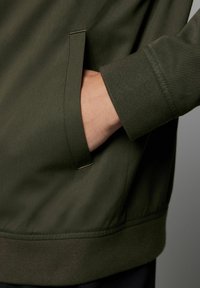 Olivgrüne Jacke aus strukturiertem Stoff, mit einer Seitentasche und gerippten Bündchen. Das Design ist modern und praktisch, mit klaren Linien.