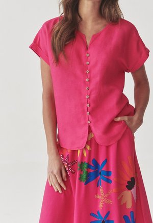 Femme portant une chemise rose vif à manches courtes boutonnée et une jupe assortie avec de grands motifs floraux colorés.