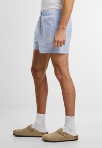 Shorts rayés bleus et blancs avec une taille élastique, associés à des sabots beiges et des chaussettes blanches. Les shorts présentent un emblème décoratif.