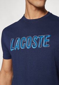 Navy modra bombažna majica s poudarjenim večbarvnim grafičnim logotipom "LACOSTE" v gradientnih modrih odtenkih čez prsi.