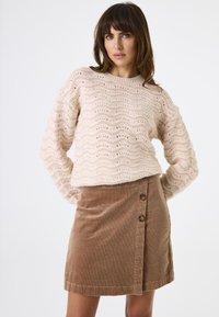 Lichtroze gebreide trui met golvend patroon en lange mouwen, gecombineerd met een bruine corduroy rok met een zijsluiting met knopen.