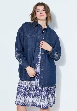 Frau trägt eine dunkelblaue bestickte Jeansjacke über einem weiß-blau gemusterten knielangen Kleid und steht vor weißem Hintergrund.