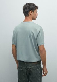 Man med kort mörkt hår som bär en ljusgrå kortärmad skjorta och svarta jeans, stående sidledes mot en enkel ljus bakgrund.