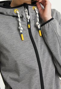 Haut à capuche zippé gris en tissu doux avec une capuche noire. Il présente des cordons blancs et jaunes et une fermeture éclair noire.