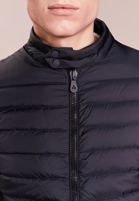 Tmavě modrá péřová bunda s stojatým límcem, opatřená předním zipem a dvěma zapínacími knoflíky, prošívaná textura a hladký povrch tkaniny.