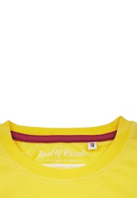 Keltainen puuvillapaita, jossa on pyöreä kaula-aukko. Paidan reunassa on viininpunainen kaulustrimmi ja sisäetiketti, joka kertoo brändistä ja alkuperästä.