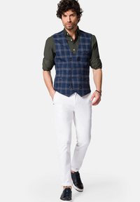 Granatowa kamizelka w kratkę noszona na ciemnozielonej koszuli, w zestawie z białymi spodniami i granatowymi trampekami. Smukły krój z zapięciem na guziki i podwiniętymi rękawami.