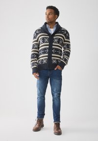 Marineblauer und cremefarbener gemusterter Cardigan mit Schalkragen, Knöpfen und gerippten Bündchen, kombiniert mit blauen Jeans und braunen Schnürstiefeln.
