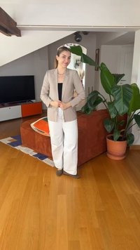 Νεαρή γυναίκα με μπεζ σακάκι, μαύρο τοπ και άσπρο παντελόνι στέκεται χαμογελαστή σε ένα μοντέρνο σαλόνι με μεγάλο πράσινο φυτό και σκουριασμένο καναπέ.