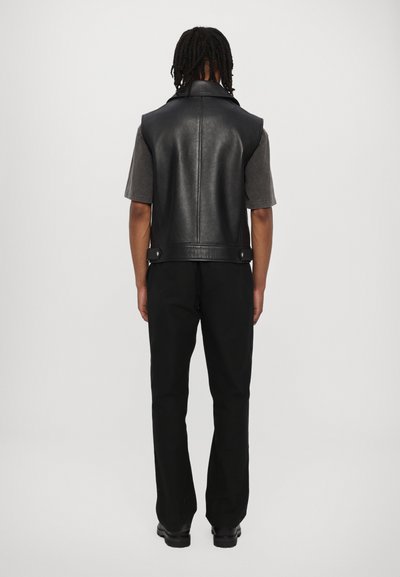 Zwarte leren vest met een hoge kraag, rugpand en drukknoopsluitingen. Gepaard met een grijs overhemd met korte Mouwen en zwarte broek.