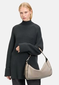 Ciemnozielony, dziany sweter z wysokim dekoltem i luźnym krojem. Modelka trzyma beżową skórzaną torbę o zakrzywionym kształcie i krótkim pasku.