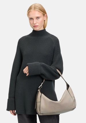 Dunkelgrüner, gestrickter Pullover mit hohem Kragen und oversize Passform. Das Model hält eine beige Lederhandtasche mit geschwungener Form und kurzem Trageriemen.