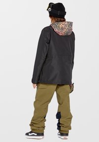 Czarna wodoodporna kurtka z kolorowym kapturem w panterkę, zestawiona z oliwkowymi szerokimi spodniami i czarnymi butami do skateboardu.