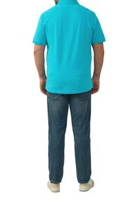 Tropfenshirt in Türkis, kurze Ärmel, glatte Textur, getragen mit blauen Jeans und hellen Schuhen. Von hinten gesehen.