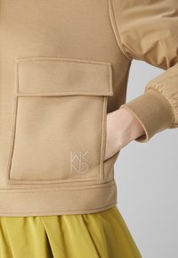 Beżowy sweter z dużą kieszenią z przodu, prążkowanymi mankietami i logo. Materiał jest gładki z delikatną teksturą, zestawiony z żółtym plisowanym dołem.