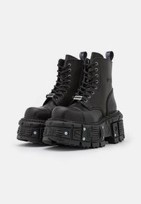 Svarta ankelskor i slät läder med tjocka, texturerade plattformssålar. Har snörning framtill och en dragflik baktill för enkel påtagning.