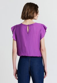 Donna che indossa una blusa corta a maniche corte di colore viola con una piccola apertura a bottone sul retro, abbinata a jeans blu scuro a vita alta.