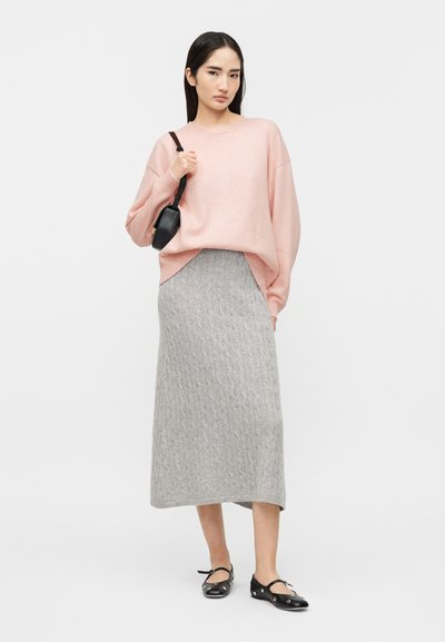 Νεαρή γυναίκα που φοράει ένα ροζ φαρδύ πουλόβερ, γκρι midi φούστα με πλεκτό σχέδιο κοτσίδα, μαύρα μπαλαρίνια με λουράκια και κρατά μια μικρή μαύρη τσάντα ώμου.