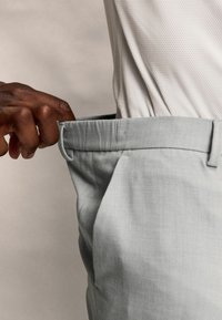 Pantalones de vestir gris claro con una textura suave, que presentan una cintura ajustada y un bolsillo lateral. Una mano ajusta la cintura.