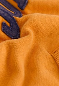 Tela naranja que muestra una textura suave con fibras visibles. Un detalle bordado de color azul oscuro aparece en la esquina superior izquierda.