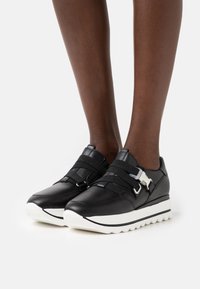 Baskets en cuir noir avec des sangles élastiques et des éléments en métal argenté, dotées d'une semelle en caoutchouc blanc épais et d'un design épuré et arrondi.