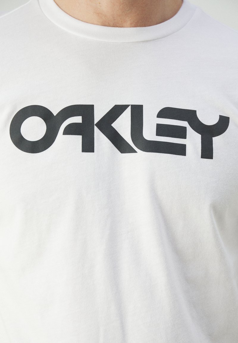 Camiseta de algodón blanca con un gran logo negro "OAKLEY" en una fuente moderna en el pecho. Diseño clásico de cuello redondo.