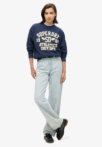Sweatshirt marine avec texte graphique blanc, coupe décontractée ; jeans larges bleu clair ; chaussures noires ; modèle se tient les mains sur les hanches.