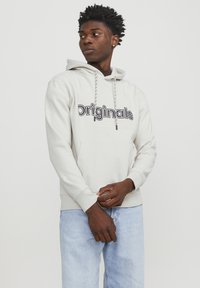 Sudadera de color beige claro con un bolsillo frontal y capucha con cordón. Presenta el logo "Originals" en texto negro en negrita. Tejido suave y casual.