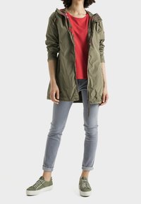 Personne portant une veste à capuche vert olive par-dessus une chemise rouge, un jean gris clair retroussé et des baskets vert olive avec des semelles blanches.