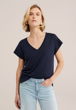 Femme portant des boucles d'oreilles anneaux argentés, un haut bleu marine à col en V manches courtes et un jean bleu clair, debout avec une main dans la poche.