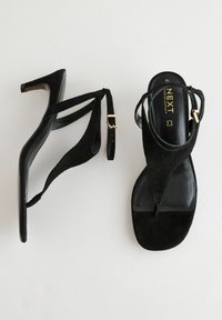 Sandali con tacco in suede neri con design a dito, caratterizzati da fasce sottili e un tacco rettangolare. Accento in oro sulla cinghia alla caviglia.