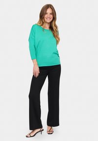 Turkio spalvos didelis megztinis su trim ketvirtadaliais rankovėmis, derinamas su juodomis plačiomis kelnėmis ir juodais šlepetėmis ant juostos. Lygus, lengvas audinys.