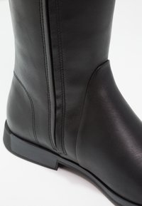 Botín de cuero negro hasta el tobillo con una textura suave, cremallera lateral y suela de goma plana. Presenta costuras sutiles y un diseño de punta redonda.