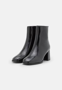 Bottes courtes en cuir noir avec une finition brillante, un bout carré et un talon épais. Texture lisse avec peu de détails de couture.
