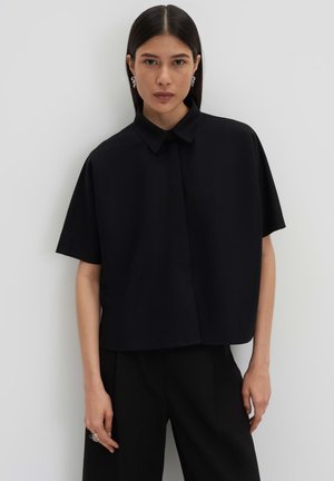 Vrouw met steil donker haar draagt een oversized zwart shirt met korte mouwen en zwarte broek, staand tegen een effen witte muur.
