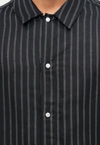 Κοντινό πλάνο μαύρου πουκάμισου με λεπτές κάθετες λευκές ρίγες και τρία ορατά λευκά κουμπιά, που δείχνει το γιακά και την άνω περιοχή του στήθους.