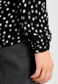 Chemise noire avec étoile blanche et motifs floraux, tissu texturé, et poignets à boutons. Portée avec un bas gris à motifs.