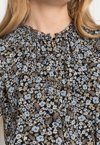 Schwarze Bluse mit einem floralen Muster in Blau, Beige und Weiß, mit gerafftem Ausschnitt, Knopfverschluss und texturiertem Stoff.