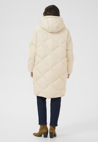 Beige puffjakke med diamantquiltet mønster, hætte og lange ærmer, parret med mørkeblå jeans og brune støvler med hæl.