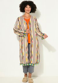 Wielokolorowy kardigan w stylu kimono z pionowymi paskami, fr�ędzlami na dole i długimi rękawami. Noszony na pomarańczowej bluzce i szarych, skróconych spodniach.