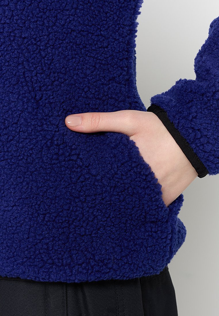 Mano inserita nella tasca di una giacca in fleece blu con polsini neri, mostrata dalla vita al polso su sfondo neutro.