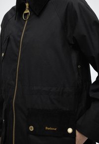 Gros plan d'une veste Barbour noire avec une fermeture éclair dorée, un col en velours côtelé et un rabat de poche avant avec un logo brodé en or et des boutons-pression.