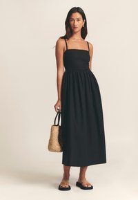 Vestido largo negro con corsé ajustado y cintura elástica, con tirantes finos y falda fluida. Bolsa tejida y sandalias planas.