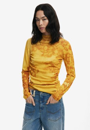 Gelbes Langarmoberteil mit floralen Mustern und Textur. Kombiniert mit blauen Jeans im relaxed Fit und Fünf-Taschen-Design.