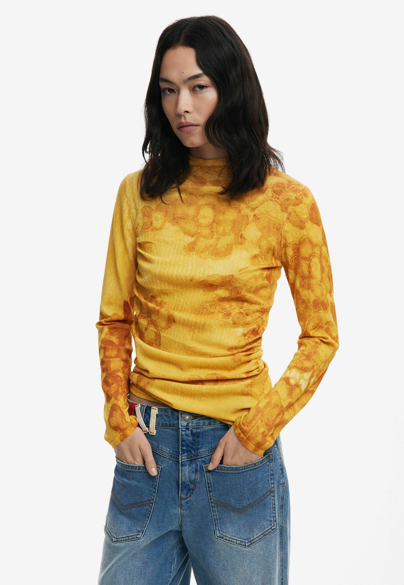 Gelbes Langarmoberteil mit floralen Mustern und Textur. Kombiniert mit blauen Jeans im relaxed Fit und Fünf-Taschen-Design.