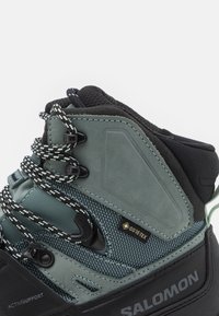 Grønn og svart fjellstøvel i middehøyde med teksturert semsket skinn og mesh, med et Gore-Tex-merke og mønstrede lisser for en sikker passform.